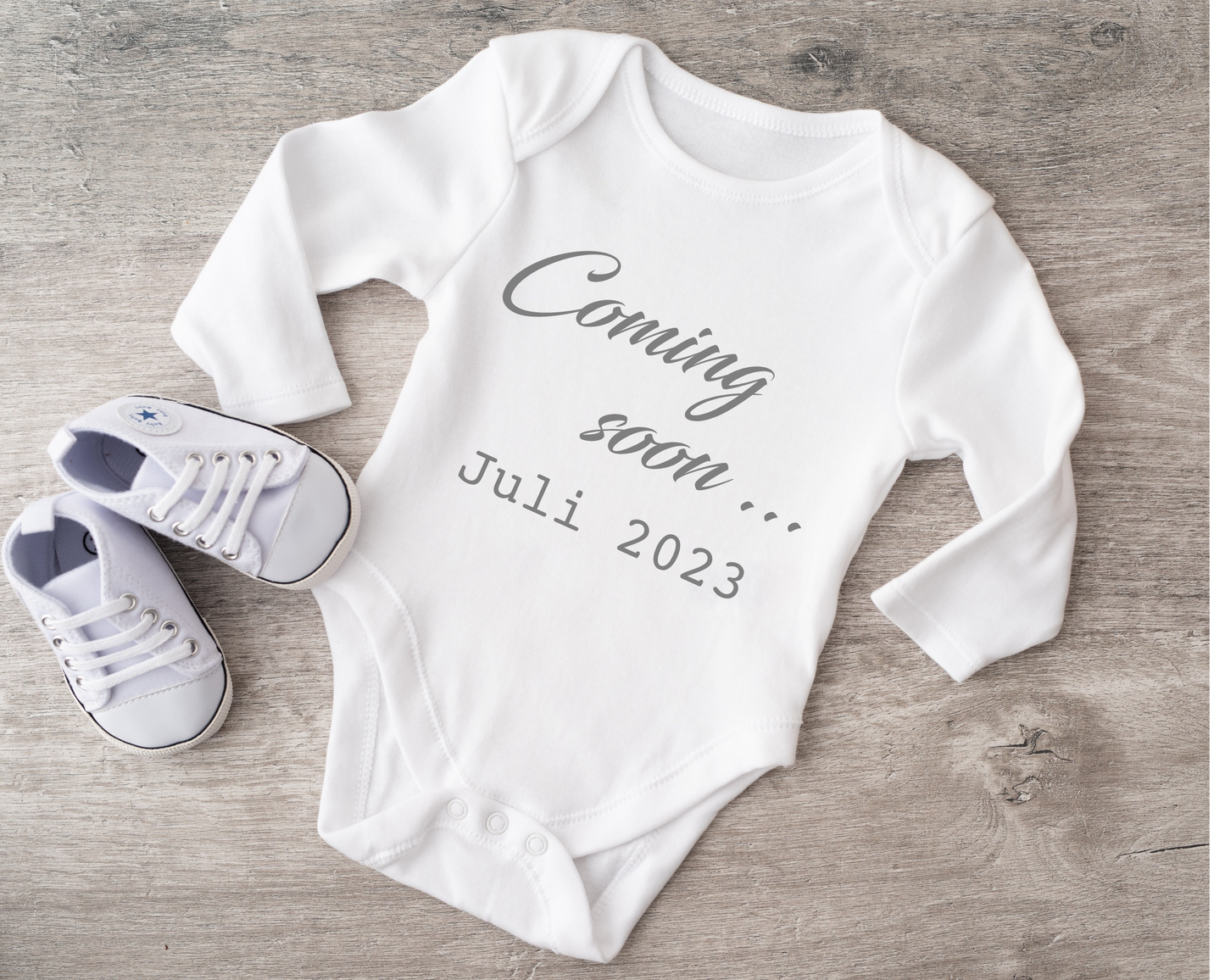 White baby bodysuit with "Coming soon... Juli 2023" text, beside baby shoes on wooden surface.