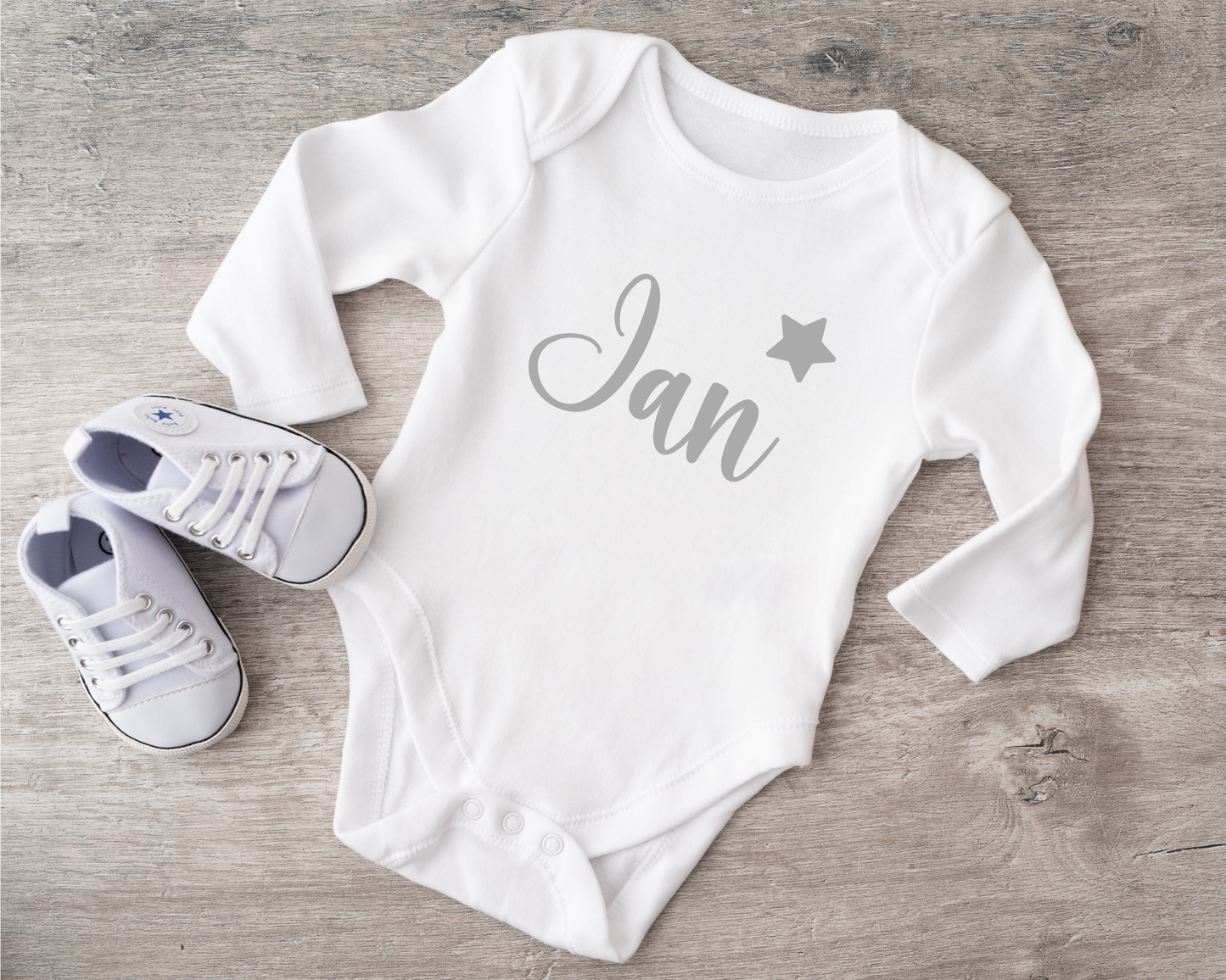 White babybody with "Jan" text and star design, paired with baby shoes, on wooden surface.