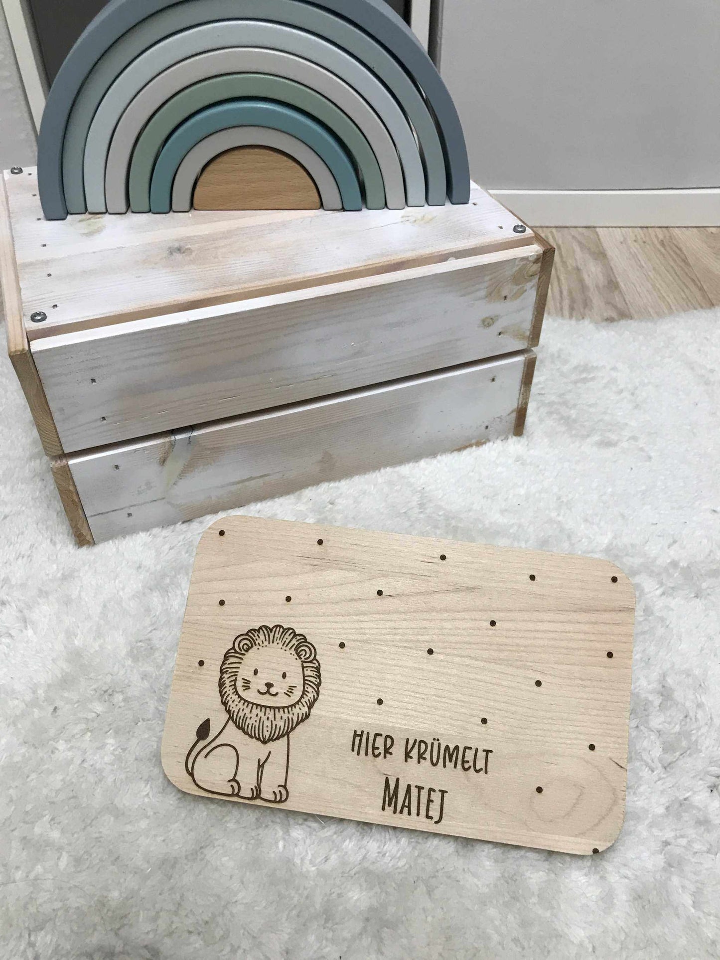 Löwe Frühstücksbrettchen aus Holz mit personalisierter Gravur und Punktdesign, auf hellem Teppich neben Spielzeug-Regenbogen.
