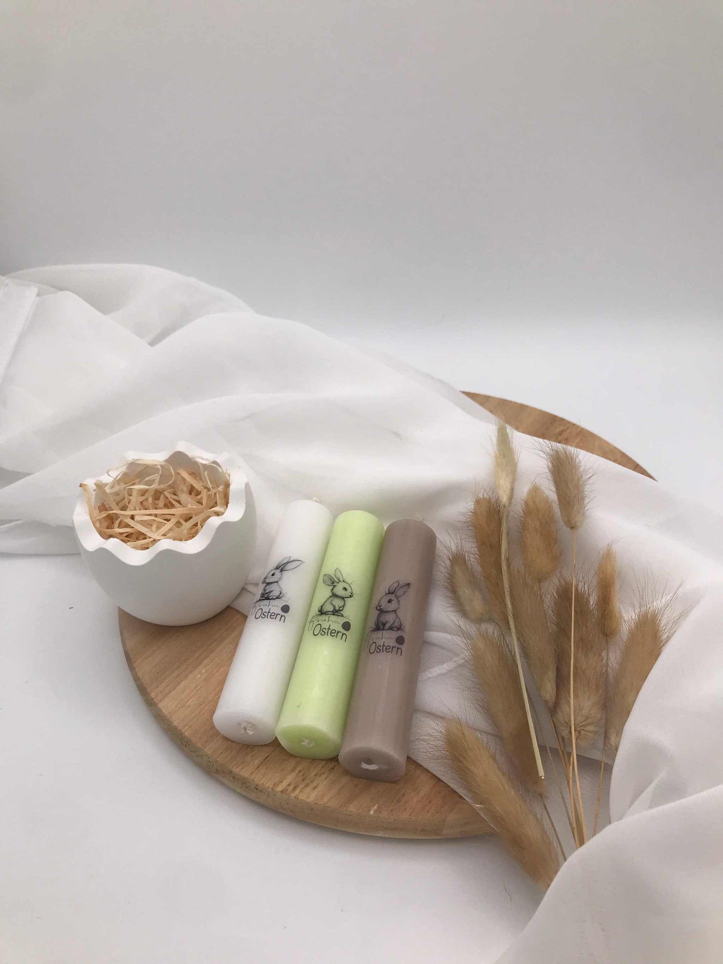 Oster-Stabkerzen mit Hasenmotiv in Pastellfarben auf Holzunterlage, dekoriert mit Trockenblumen und weißem Tuch.