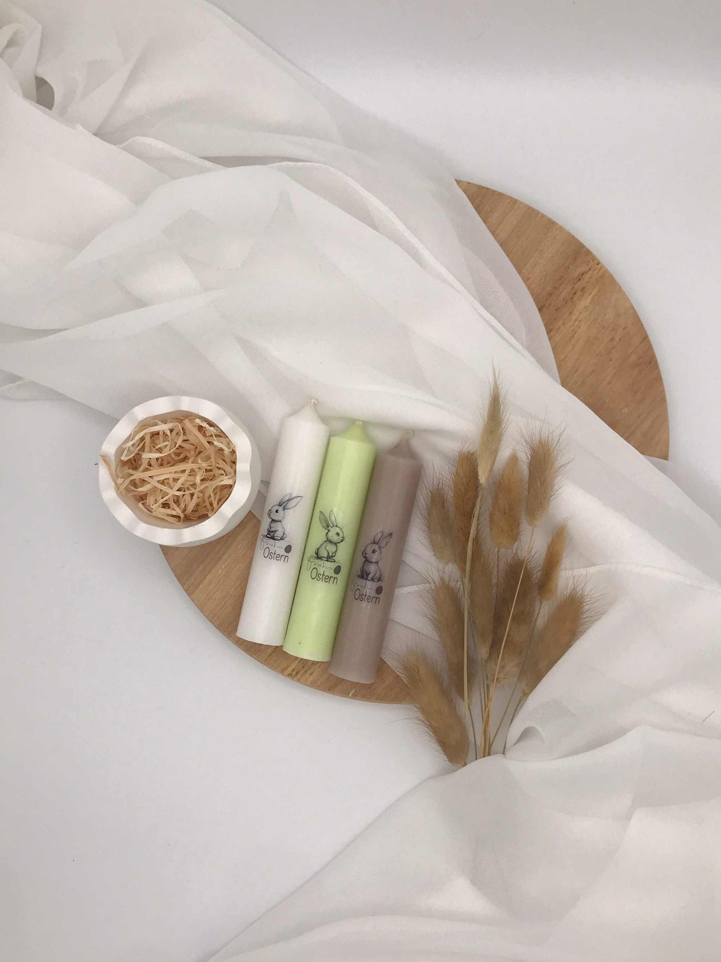 Oster-Stabkerzen mit zarten Hasenmotiven in Pastellfarben auf einem Holztablett, dekoriert mit Gräsern.