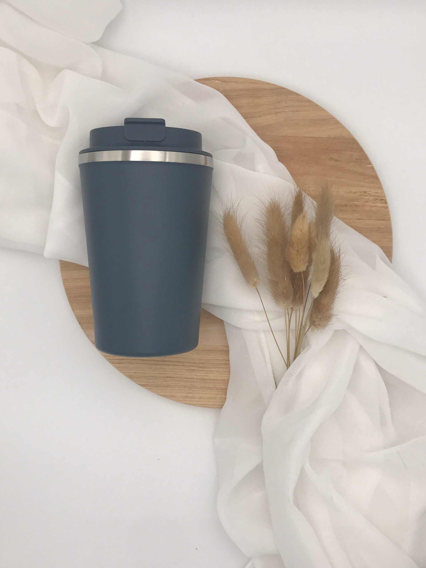 Doppelwandiger Edelstahlbecher mit Gravur auf einem Holztablett, umgeben von weißem Stoff und Trockenblumen.