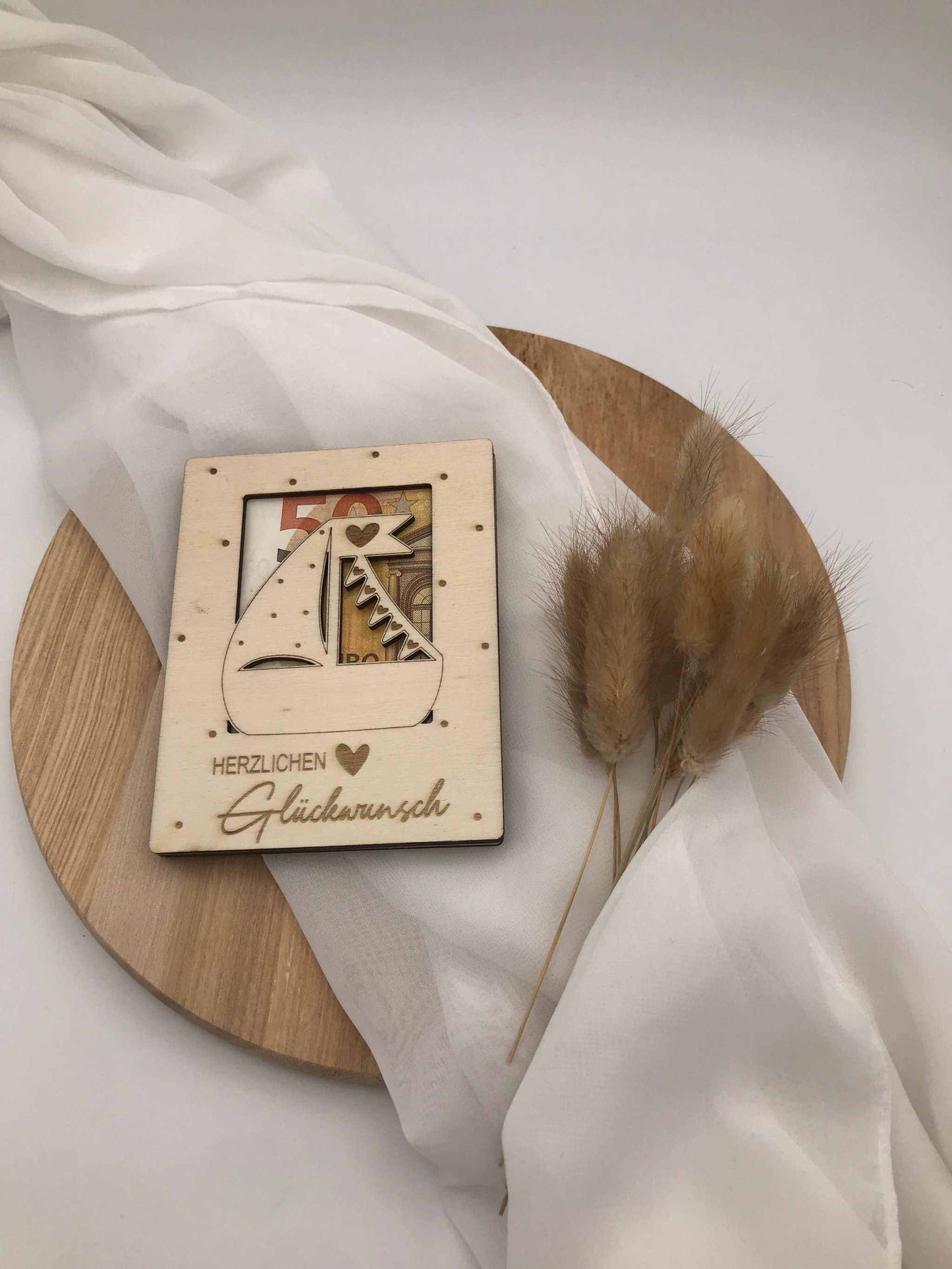 Geldgeschenk aus Sperrholz zur Taufe, Kommunion, Firmung oder Konfirmation, dekorativ auf Holzplatte mit Trockenblumen und Tuch arrangiert.