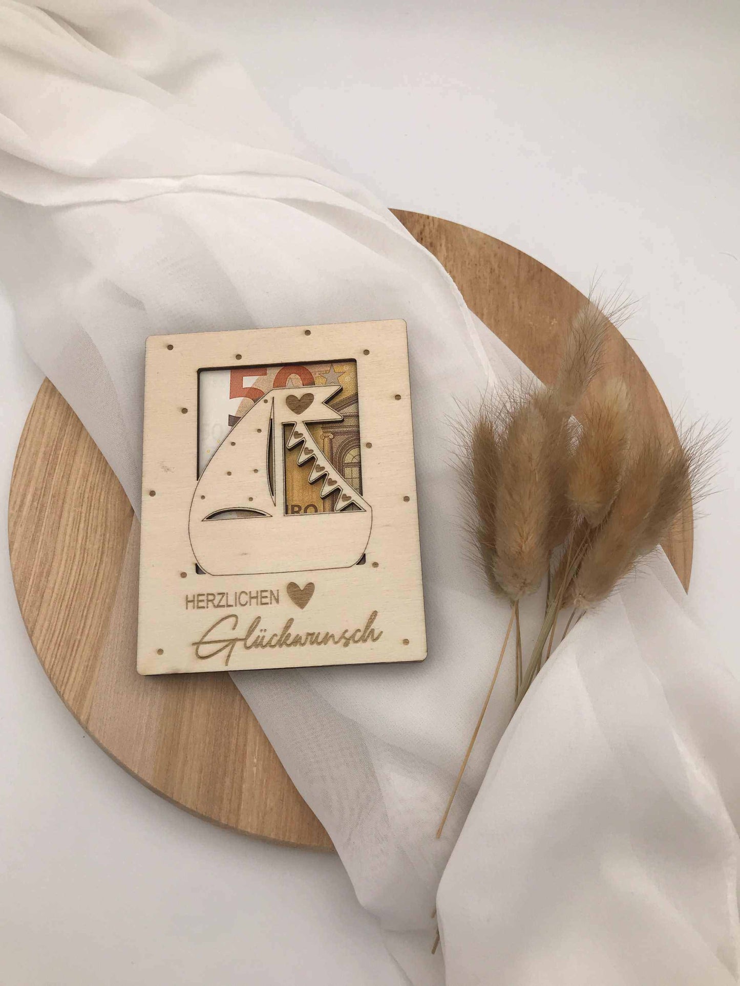 Geldgeschenk zur Taufe aus Holz mit eingraviertem Segelboot-Design auf Holzplatte, dekoriert mit Trockenblumen.