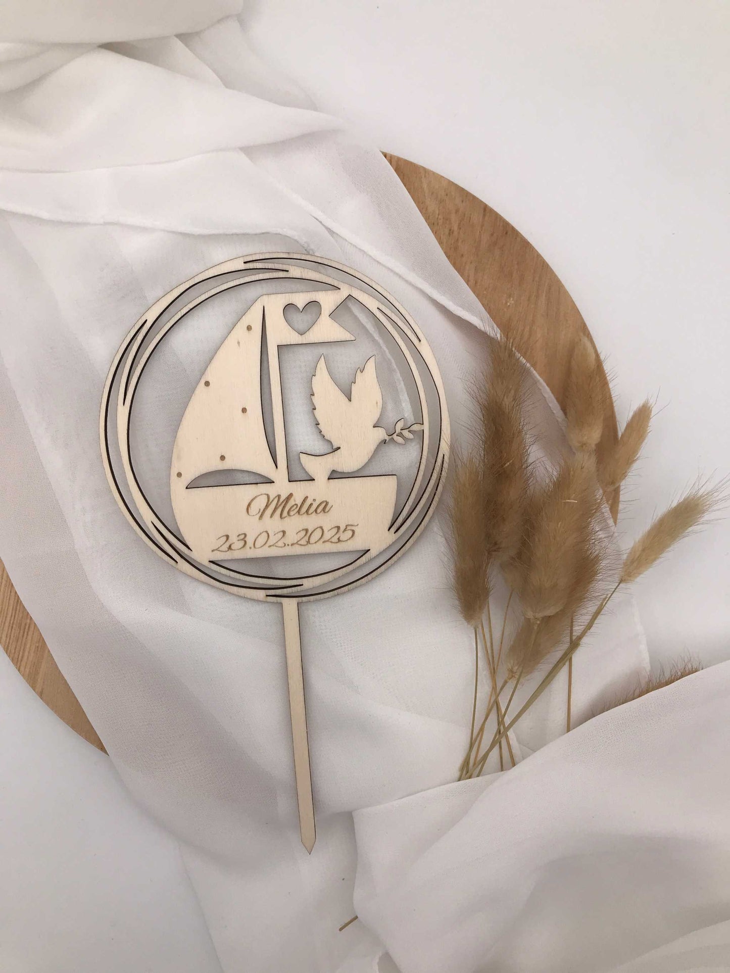 Caketopper aus Holz mit personalisiertem Design zur Taufe, daneben liegen getrocknete Gräser auf weißem Tuch.