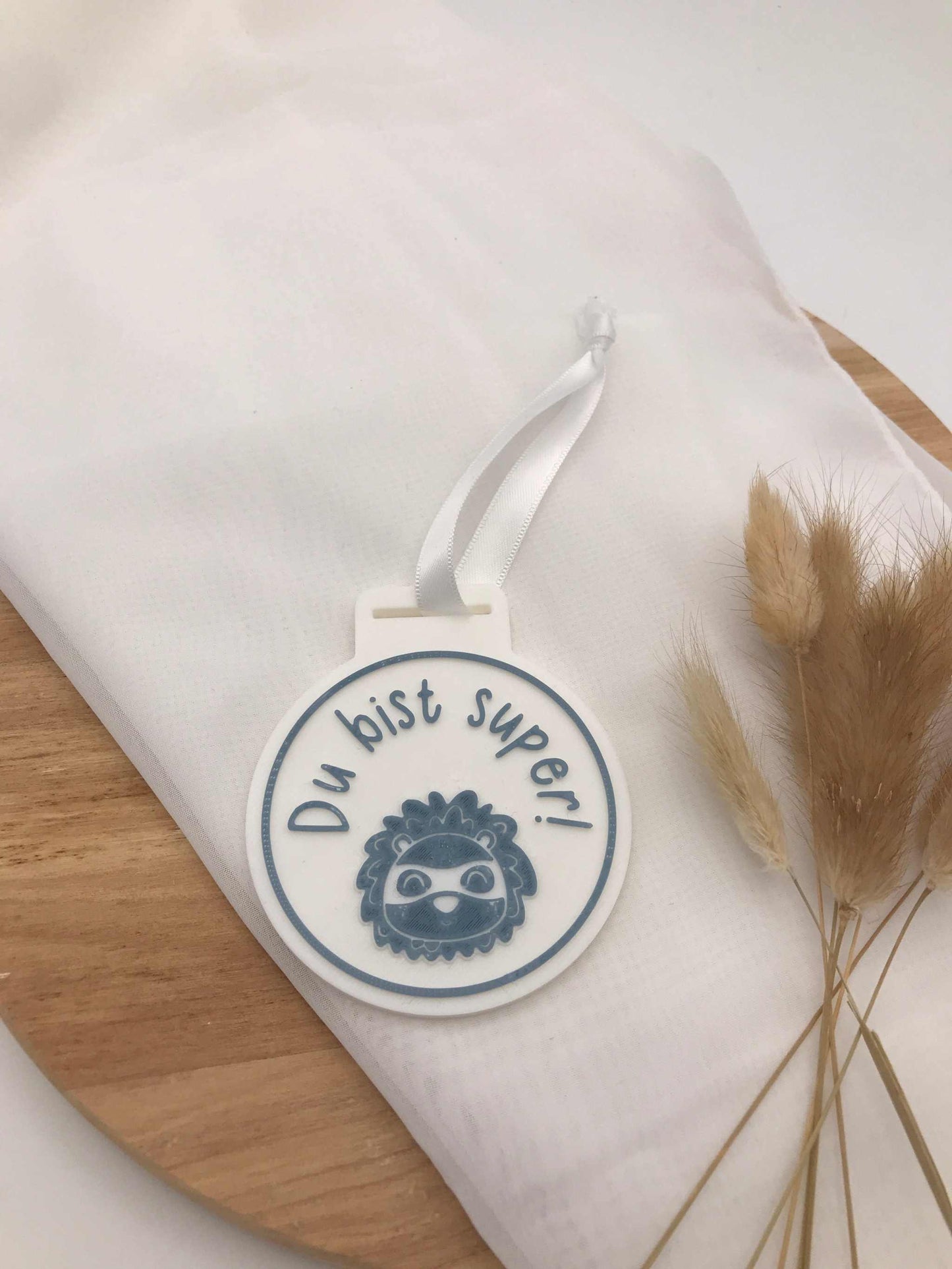weiße 3D-Druck Medaille zur Einschulung mit dem Spruch "Du bist super" und Riemen auf Stoff und Holz mit Trockenblumen