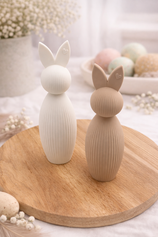 Zwei moderne Osterhasen aus PLA in Creme und Beige auf einem runden Holzbrett, minimalistische Frühlingsdekoration