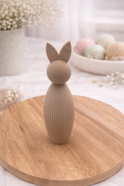 Beiger Osterhase aus PLA mit geriffelter Oberfläche auf rundem Holzbrett, frühlingshafte Deko mit pastellfarbenen Ostereiern im Hintergrund