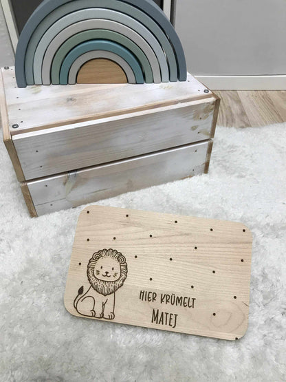 Löwe Frühstücksbrettchen aus Holz mit personalisierter Gravur und Punktdesign, auf hellem Teppich neben Spielzeug-Regenbogen.