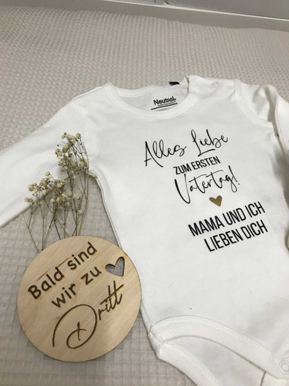 weißer Babybody mit Aufdruck "Alles Liebe zum ersten Vatertag! Mama und ich lieben dich" und Holzschild "Bald sind wir zu dritt" mit getrockneten Blumen auf Stoffuntergrund