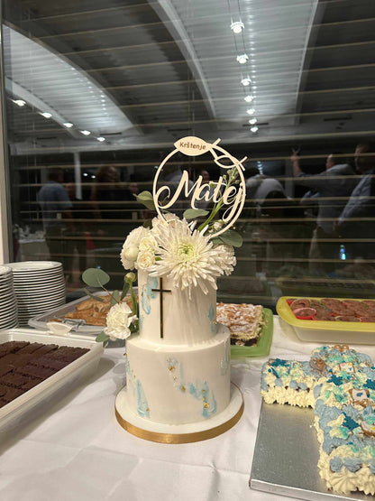 Caketopper aus Holz auf weißer zweistufiger Torte mit Blumendekoration bei Feier im Innenbereich.