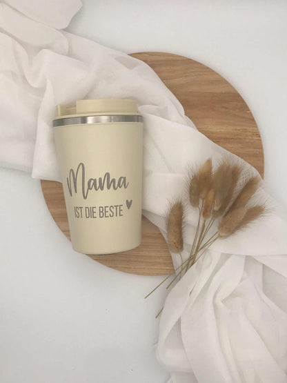 Doppelwandiger Edelstahlbecher mit Gravur "Mama ist die Beste" auf Holzuntergrund und weißem Stoff.