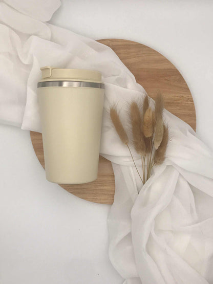 Doppelwandiger Edelstahl-Thermobecher in cremefarben auf Holzbrett mit weißem Stoff und Trockenblumen dekoriert.