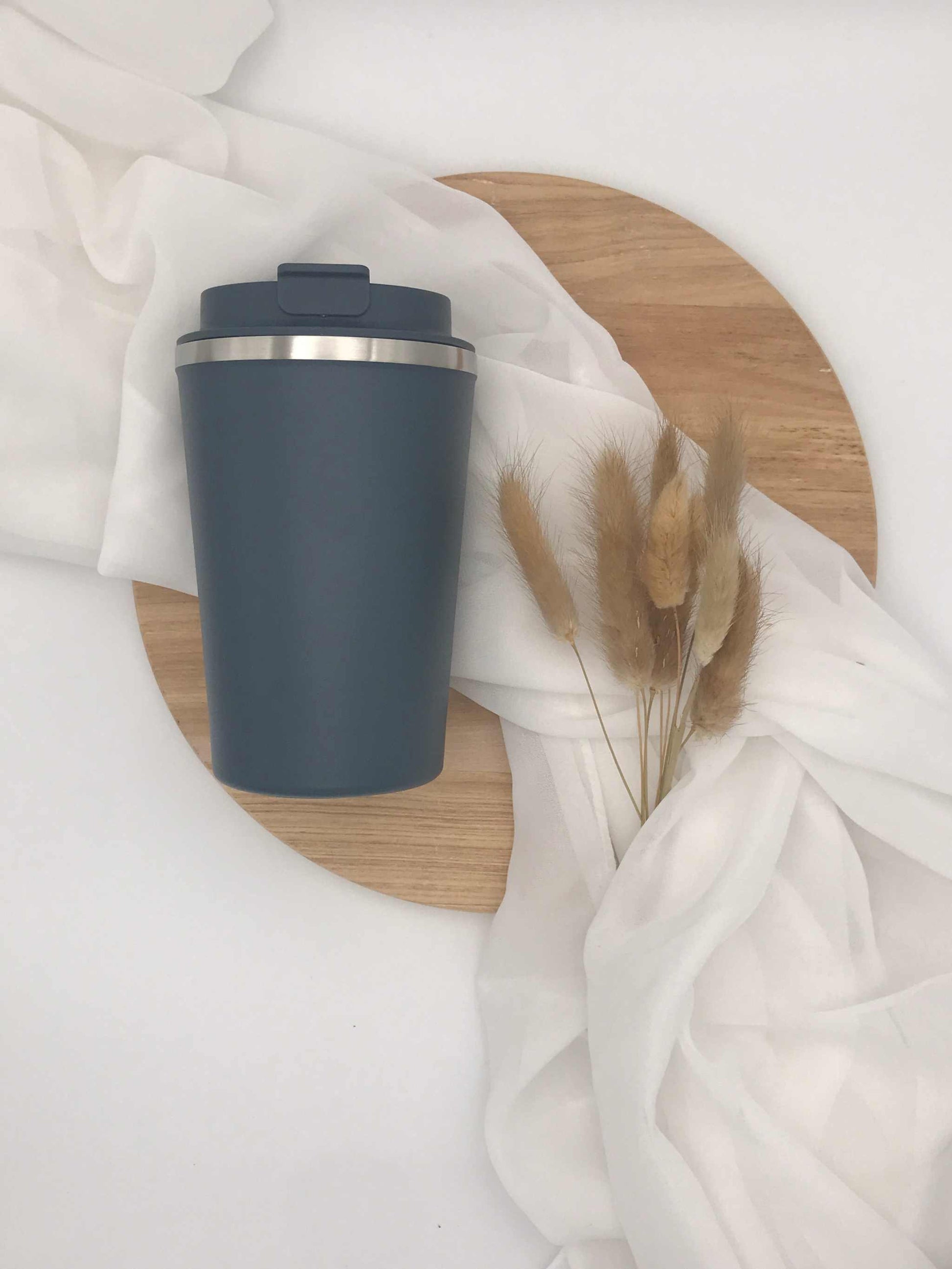 Doppelwandiger Edelstahlbecher mit Gravur auf einem Holztablett, umgeben von weißem Stoff und Trockenblumen.