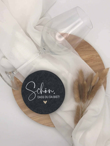 Felt coaster set with "schön, dass du da bist" design, anthracite color, on a wooden background with a glass.