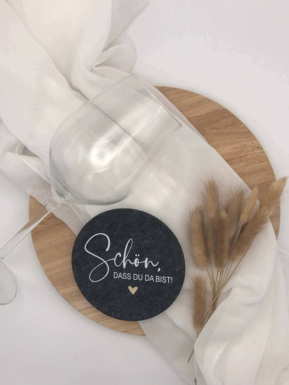 Filzuntersetzer mit Schriftzug "Schön, dass du da bist" auf Tisch, dekoriert mit Glas und weißen Stoffservietten.