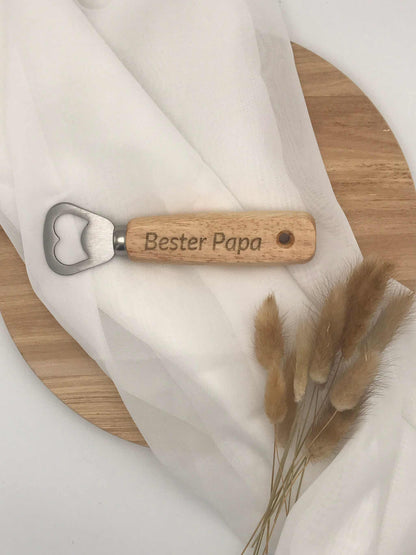 Wooden bottle opener with "Bester Papa" engraving on oak handle, stainless steel opener, on a light wooden surface with fabric and dried flowers.
