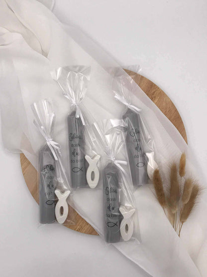 Gastgeschenke zur Taufe und Kommunion, graue Kerzen mit Fischsymbol verpackt auf Holzplatte.