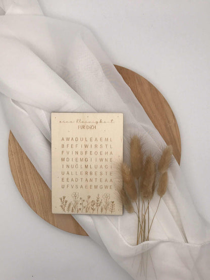 Schwangerschaftsverkündung als Worträtsel auf Holzkarte mit elegantem Design, auf hellem Hintergrund mit Trockenblumen.