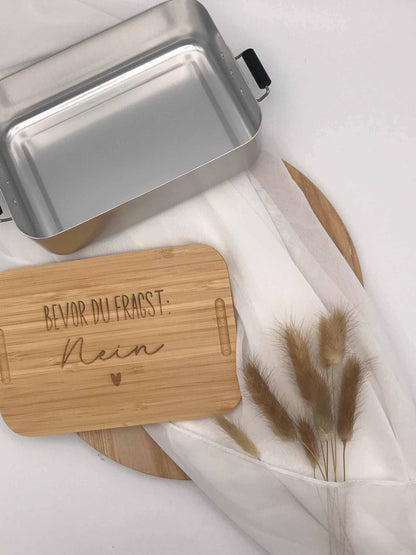 Brotdose mit Bambusdeckel und Gravur auf weißem Hintergrund, geöffnet, neben Trockenblumen.