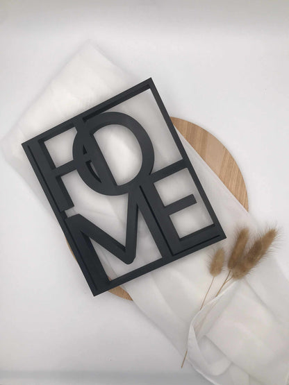 3D-Druckschriftzug „Home“, schwarz, auf weißem Tuch und Holzunterlage, stilvolle Wohnaccessoire-Dekoration.