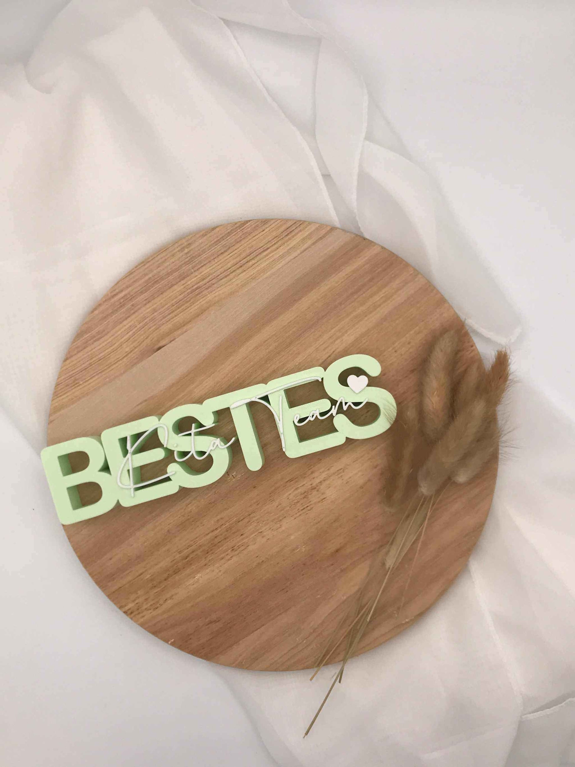 3D-Druck Aufsteller "Bestes Kita-Team" aus Holz auf weißem Stoff, dekorativ mit Trockenblumen arrangiert.