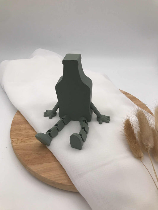 3D-printed green decoration beer bottle with movable wobbly legs sitting on white cloth and wooden board