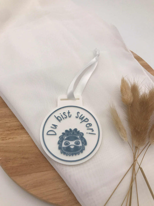 weiße 3D-Druck Medaille zur Einschulung mit dem Spruch "Du bist super" und Riemen auf Stoff und Holz mit Trockenblumen