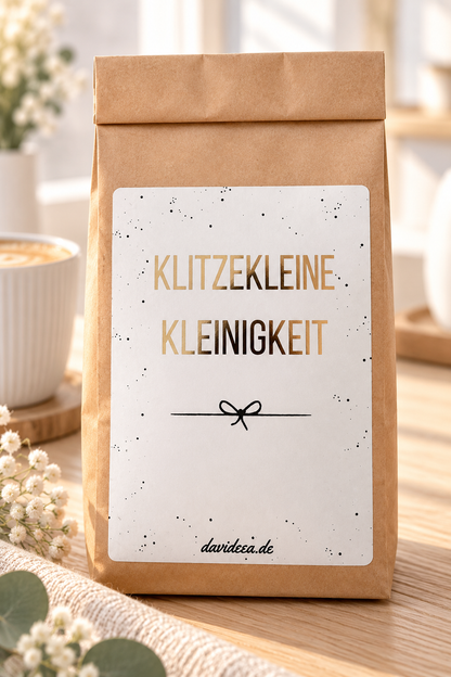 Geschenktüte als kleine Aufmerksamkeit aus braunem Kraftpapier mit weißem Etikett, goldener Schrift „Klitzekleine Kleinigkeit“ und schwarzer Schleife