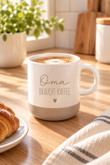 Tasse aus cremefarbener Keramik mit beigem Boden, feinen Sprenkeln und dem Aufdruck „Oma braucht Kaffee“ samt Herz