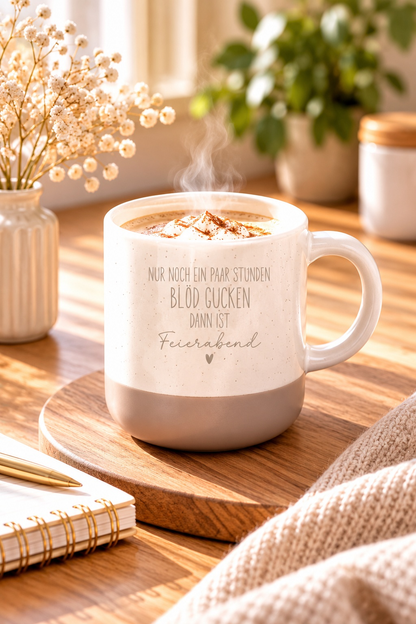 Tasse aus cremefarbener Keramik mit beigefarbenem Fuß, feinen Sprenkeln und dem Spruch „Nur noch ein paar Stunden blöd gucken, dann ist Feierabend“.